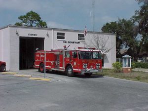 White fire station building with trees in the background and one red fire engine parked in the driveway