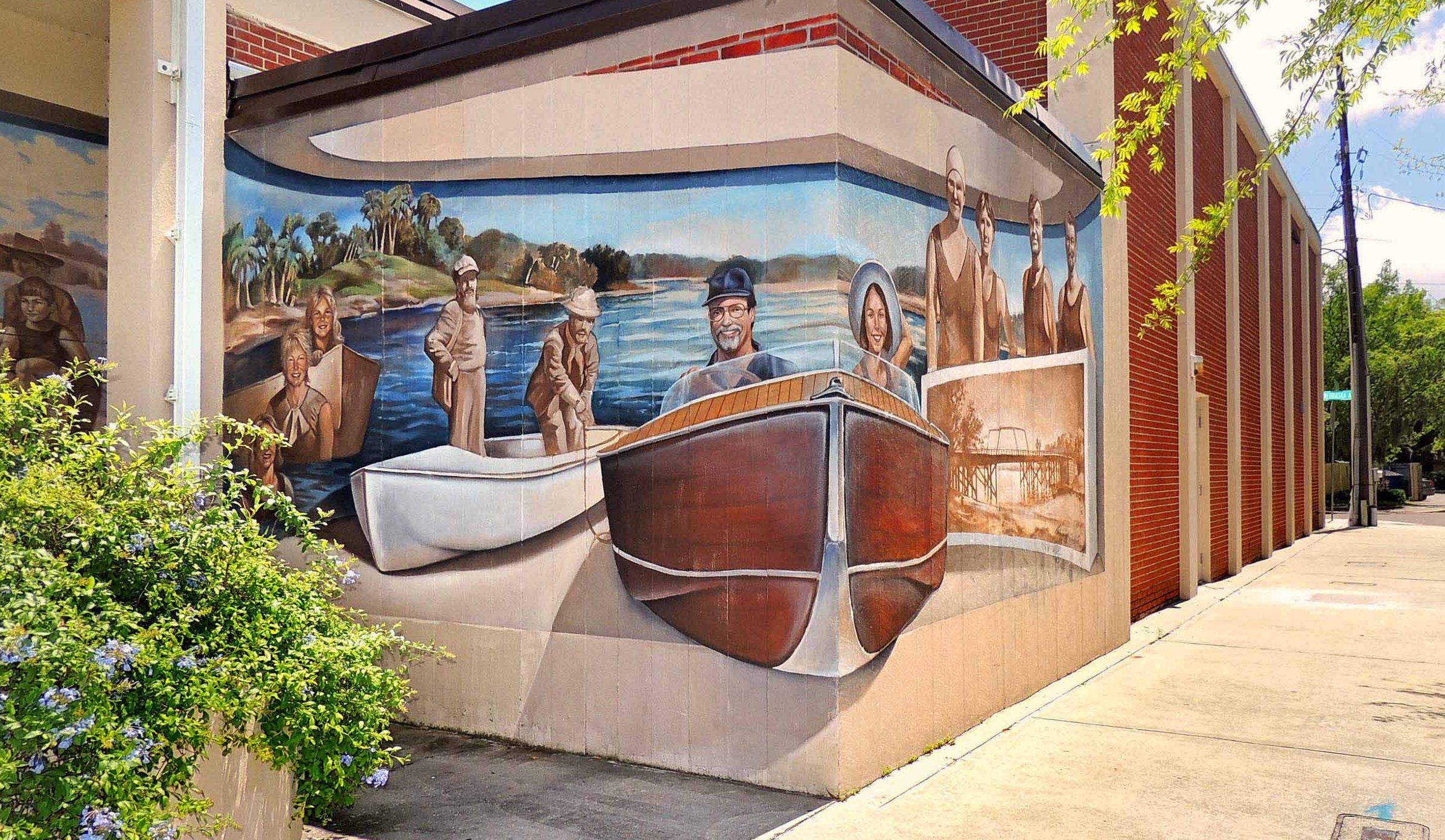 Corner of a building with a mural of happy boaters painted on it. A sidewalk stretches off into the distance.